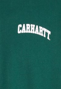 Tmavě zelená látka se znakem "CARHARTT" vytištěným tučnými bílými písmeny ve velkých všeobecně a mírně zakřivených písmenech.