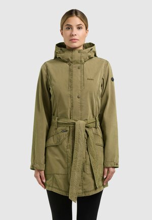 Žena v olivovo zelenej kapucňovej bunde s prednými vreckami a opaskom na zaväzovanie v páse, stojaca pred jednoduchým svetlým pozadím.