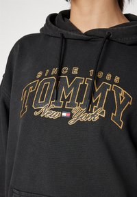 Hanorac negru cu buzunar frontal, având text galben și alb: "TOMMY" cu "New York" sub acesta, material texturat și glugă cu șnur.