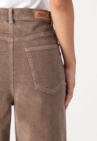Bruine corduroypantalon met een hoge taille, smalle ribstructuur en een achterzak. Een geperst leren label is zichtbaar op de tailleband.