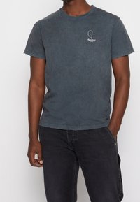 Tmavě modré tričko s krátkým rukávem vyrobené z měkké bavlny. Má opotřebený výstřih a malé bílé logo na hrudi.