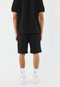 Camisa de manga corta negra texturizada y pantalones cortos a juego, combinados con zapatillas blancas y calcetines. El diseño es casual y de corte relajado.