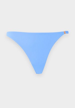 Frau steht barfuß mit einer Hand auf der Hüfte, trägt einen hellblauen Bikini vor einem schlichten weißen Hintergrund.