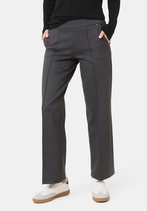 Osoba ubrana w ciemnoszare spodnie o prostych nogawkach z przednimi szwami, ręce w kieszeniach, w połączeniu z białymi trampkami i czarną bluzką z długim rękawem.