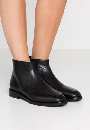 Bottines en cuir noir à talons bas portées par une personne debout sur un sol clair devant un fond uni.