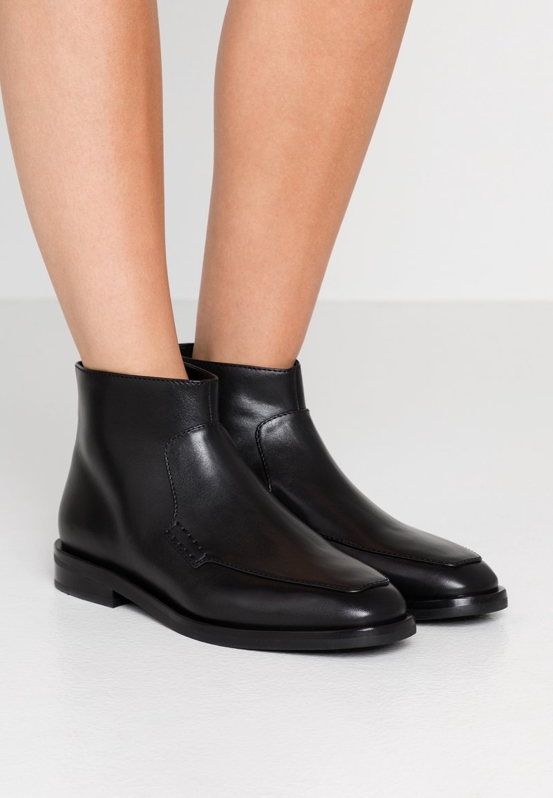 Bottines en cuir noir à talons bas portées par une personne debout sur un sol clair devant un fond uni.