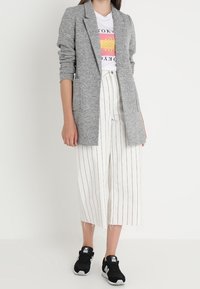 Grijze gestructureerde lange blazer, witte gestreepte wijde broek en zwarte sportschoenen. Grafisch t-shirt met roze en gele design.