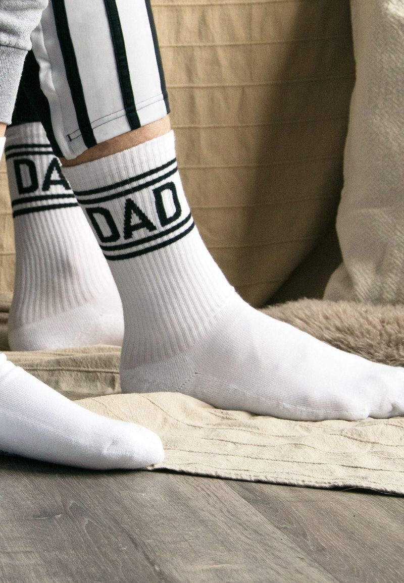 Füße, die weiße Socken mit schwarzen Streifen und dem Wort "DAD" tragen, stehen auf einem hellen Teppich über einem Holzboden.