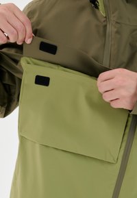 Vandtæt jakke med olivengrøn ydre stof. Har en frontlomme, der er sikret med sort velcro, og en glat, mat tekstur.