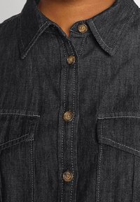 Dark denim shirt with a pointed collar, brown buttons, two chest pockets, and contrasting white stitching. Material appears sturdy and textured.