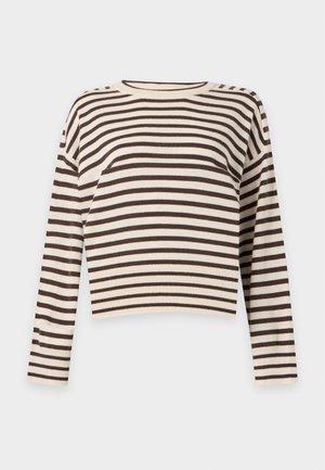 Femme portant un haut à manches longues beige et noir rayé, un pantalon large beige et des chaussures noires pointues, debout avec une main dans la poche.