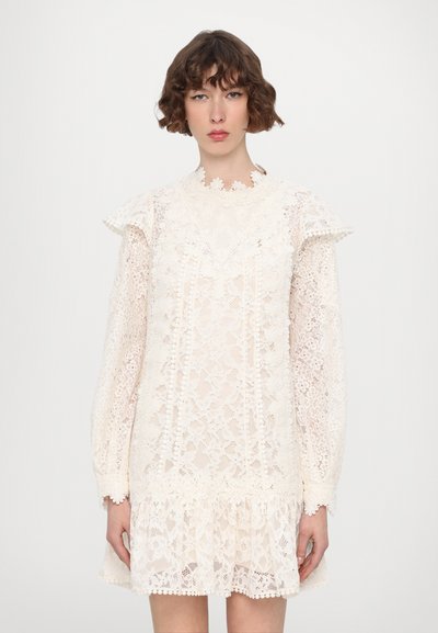 Blonde kjole i off-white med lange ærmer, rynkede skuldre og en uforholdsmæssig kant. Har blomstermønstre og bølgede kanter.