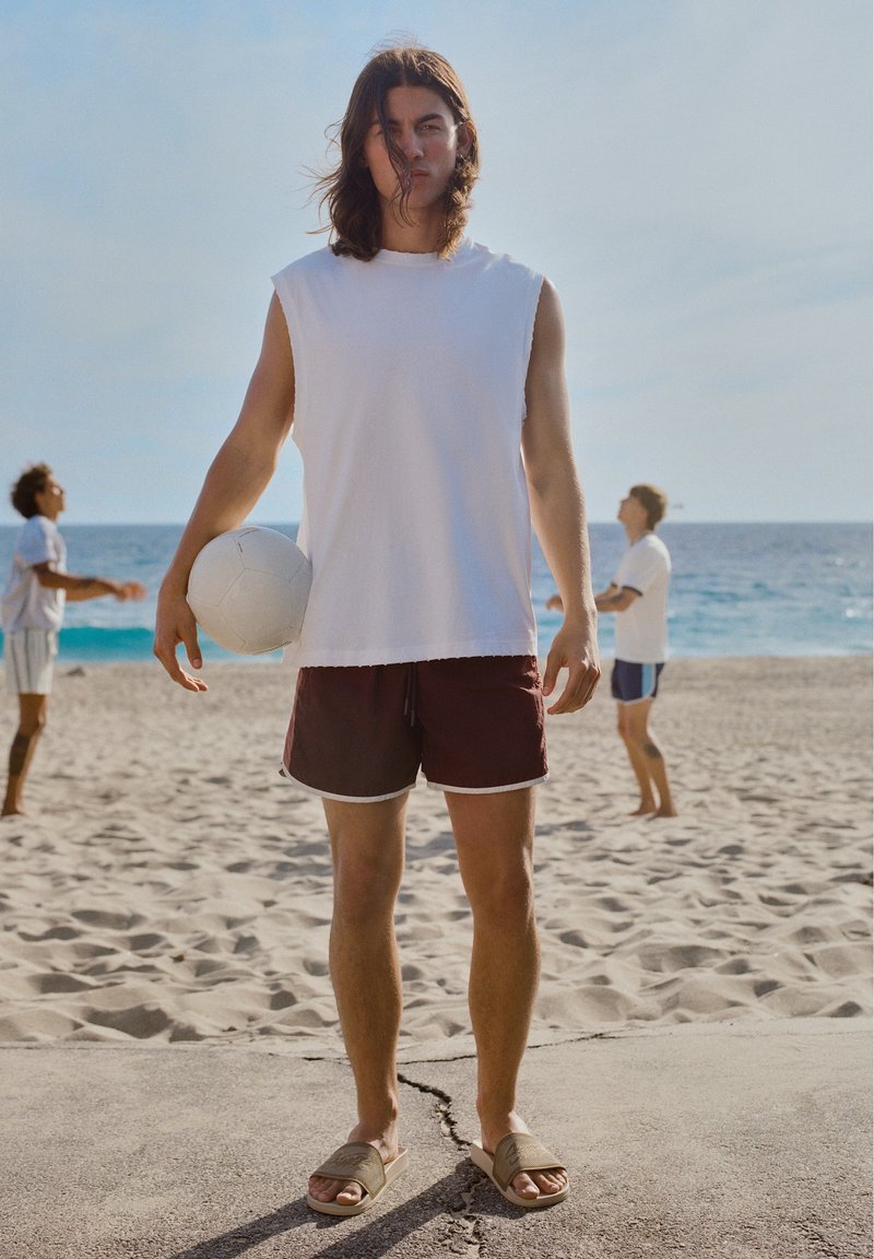 Jeune homme aux cheveux longs tient un ballon de volley-ball blanc sur la plage, portant un débardeur blanc, un short sombre et des sandales, avec deux personnes jouant derrière.