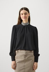 Blusa nera con maniche a sbuffo e colletto alto decorato, abbinata a una gonna di jeans beige. Tessuto liscio e dettagli plissettati visibili.