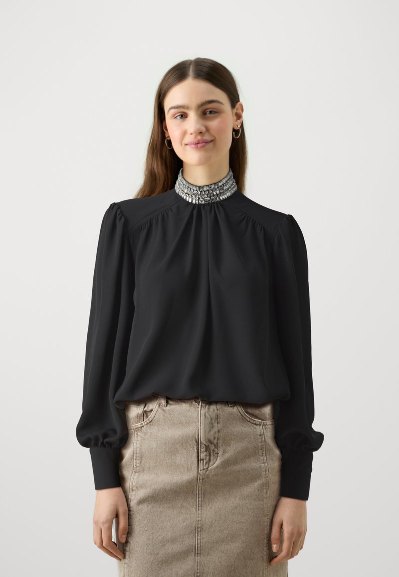 Blusa nera con maniche a sbuffo e colletto alto decorato, abbinata a una gonna di jeans beige. Tessuto liscio e dettagli plissettati visibili.
