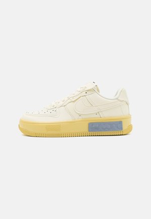 Lavtopet cremefarvet sneaker med perforeret tå, Nike swoosh, gul sål og grå rektangulær detalje på siden.