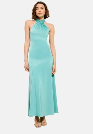 Nő ujjatlan, földig érő türkizkék nyakbakötős ruhában, kezei lazán oldalán áll egy egyszerű, világos háttér előtt.