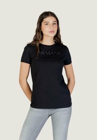 Jonge vrouw met lang bruin haar, gekleed in een zwart Armani Exchange T-shirt en lichtgrijze jeans, staand met haar handen achter haar rug.