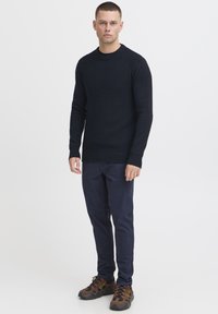 Marineblauwe geribbelde gebreide trui met een ronde hals, gedragen met donkere blauwe chinos en bruin multi-georiënteerde sneakers. Eenvoudig ontwerp, getailleerde pasvorm.