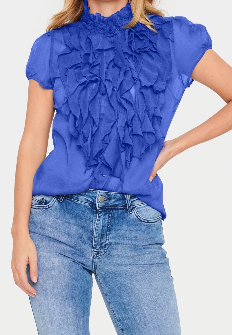 Femme portant un chemisier bleu vif à manches courtes volantées associé à un jean denim délavé clair, main posée sur la hanche.