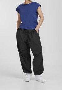 Persoon in een blauwe T-shirt met korte mouwen, losse zwarte broek met handen in de zakken, en witte sneakers, staand tegen een eenvoudige grijze achtergrond.