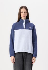 Fleece pulóver v modrej a svetlomodrej farbe s vysokým golierom a gombíkovou légou. Má textúrovaný povrch a logo GAP na hrudi.