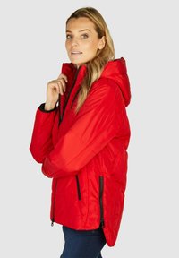 Crvena insulated puffer jakna s kapuljačom, s crnim patentnim zatvaračem i bočnim džepovima, dizajnirana s ušivenim uzorkom i lagano zakrivljenim rubom.
