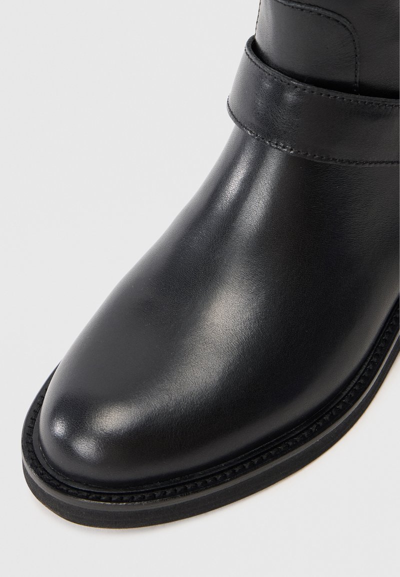 Schwarze Lederstiefelette mit abgerundeter Zehenpartie, glatter Textur und dekorativem Band am oberen Bereich. Genähte Sohlenverzierung.