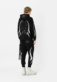 Sudadera negra con capucha y pantalones de chándal, ambos con diseños de alas y rayos blancos. El material parece suave; los detalles incluyen etiquetas y acentos. Botas blancas.
