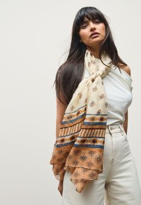 Cream and caramel scarf with intricate floral patterns, featuring blue and brown stripes and textured pleats, draped around the neck.