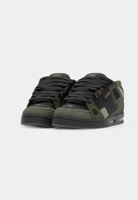 Green and black sneakers with a mix of materials, featuring a textured surface, reinforced accents, and yellow eyelets. Low-top design.