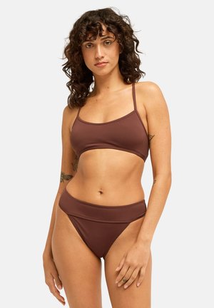 Femme aux cheveux bouclés portant un bikini marron avec des fines bretelles et un bas taille haute, debout devant un fond clair uni.