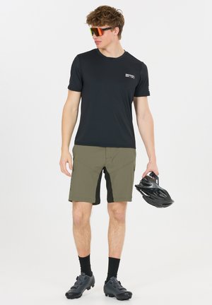 Junger Mann, der ein schwarzes Radtrikot, olivgrüne Shorts, schwarze Radschuhe und Sportsonnenbrillen trägt und einen schwarzen Fahrradhelm in der Hand hält.