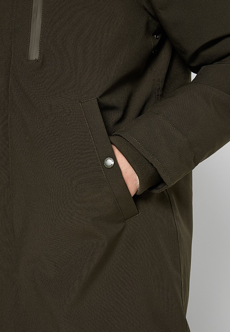 Parka imperméable vert foncé avec une poche zippée et des poches latérales. Présente une texture lisse et un accent de bouton sur la poche.