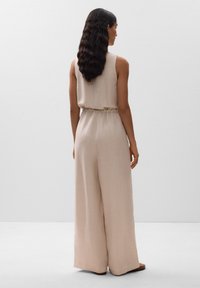 Beige Leinen-Overall mit weitem Bein, ärmellosem Oberteil, elastischem Bund und subtiler Textur, von hinten gesehen. Flache Sandalen ergänzen das Outfit.