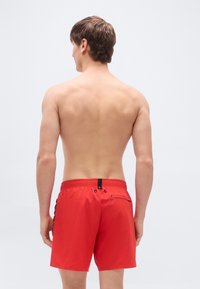 Κόκκινα μαγιό σορτς με ελαστική μέση, πίσω τσέπη και αντιθετικές μαύρες λεπτομέρειες. Λείο υφή, μήκος πάνω από το γόνατο και πλευρικές ρίγες.