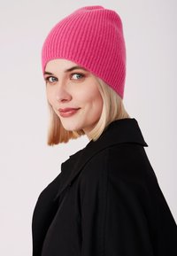 Berretto rosa lavorato a maglia con una texture a coste. Si adatta perfettamente ed ha una forma leggermente cadente, abbinato a un cappotto nero visto di profilo.