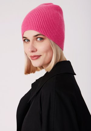 Roze gebreide muts met een geribbelde textuur. Hij zit strak en heeft een lichtjes slungelige vorm, gecombineerd met een zwarte jas in een zijaanzicht.