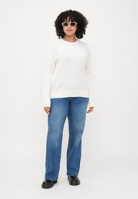Hvit, teksturert genser med langermet, kombinert med blå jeans med rett ben. Tilbehøret inkluderer lyse solbriller og svarte sko.