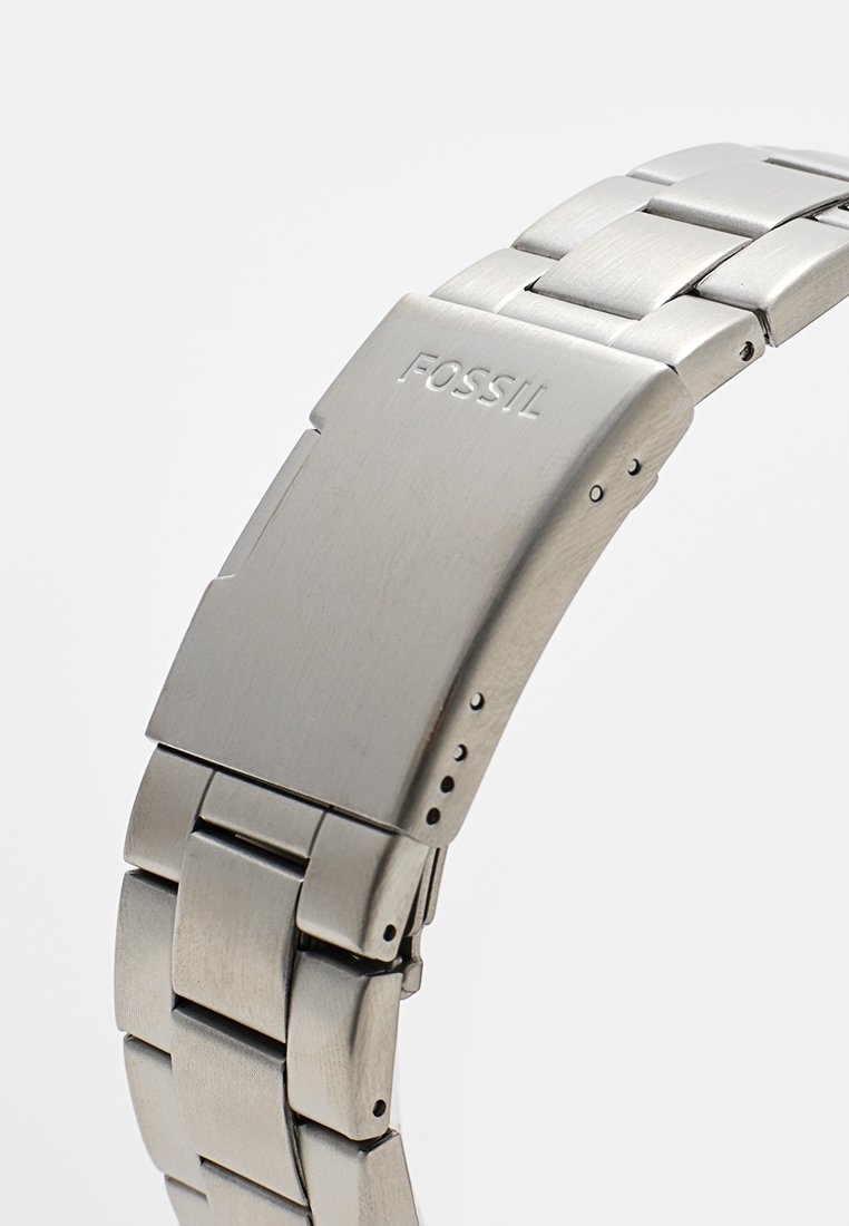 Bracciale per orologio Fossil in acciaio inossidabile con chiusura a scatto, che mostra il nome del marchio inciso e maglie regolabili su uno sfondo semplice.