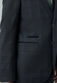 Blazer în carouri întunecate, cu un material texturat, având clapă la guler, închidere cu buton și un singur buzunar cu un accent întunecat contrastant.