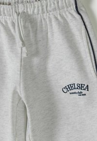 Pantalon de survêtement gris clair avec taille élastique, cordon de serrage, liseré latéral et inscription "CHELSEA tennis club est 1974" imprimée près de la poche.