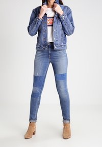 Chaqueta de mezclilla en azul medio, con cuello y cierre de botones. Camiseta blanca con logo impreso debajo, combinada con jeans azul de parche y botines beige.