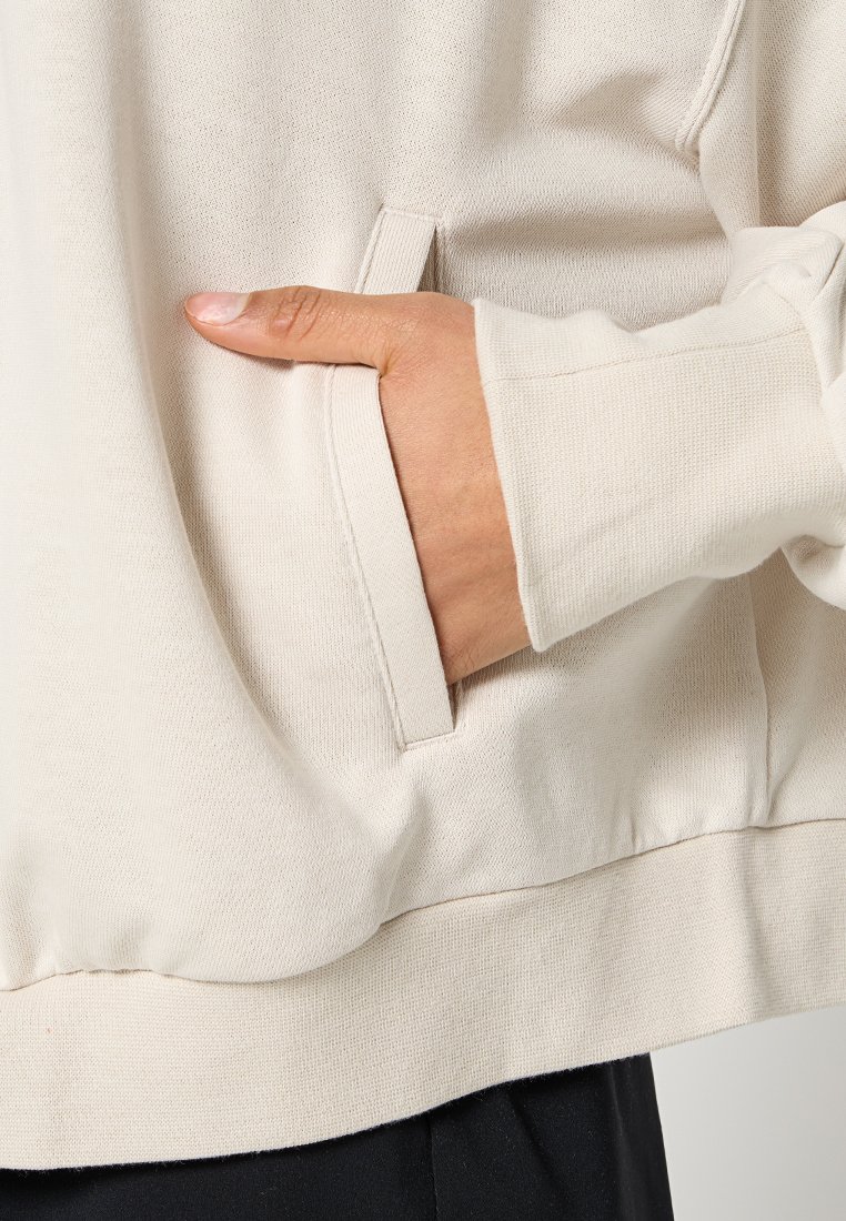 Pullover crema con lavorazione a maglia strutturata, dotato di una tasca laterale e polsini a coste, con una mano che riposa all'interno della tasca.