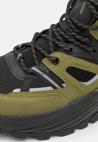 Botte de randonnée présentant un design noir et vert olive avec des matériaux en textile et caoutchouc, des superpositions texturées et des lacets durables.
