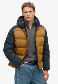 Pufferdækken med hætte, har brune og marine farveblokke, quiltet tekstur, front lynlås og justerbar hætte. Bærer over en grå sweater.