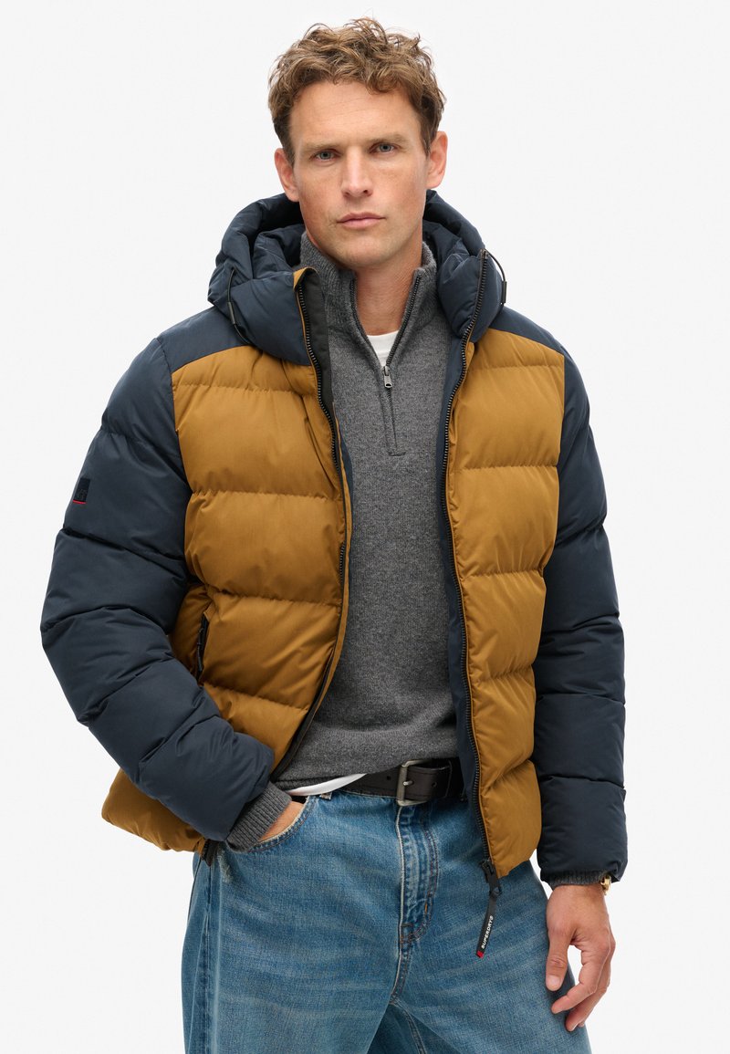 Giacca a vento con cappuccio, presenta blocchi di colore marrone e blu navy, texture trapuntata, zip frontale e cappuccio regolabile. Indossata sopra un maglione grigio.