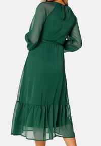 Vestido verde de longitud media con mangas largas transparentes, cintura ajustada y dobladillo con volantes. Tela texturizada con un detalle de abertura en la espalda.