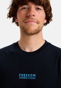 Svart bomull t-shirt med blå text som läser "FREEDOM FROM CHAOS" med en dekorativ horisontell linje, korta ärmar och rundad hals.