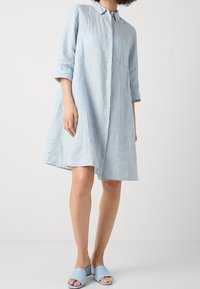  Lichtblauw gestreepte hemdjurk met knoopsluiting en kraag. Lange mouwen opgerold; gecombineerd met bijpassende blauwe instapschoenen.